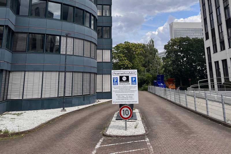 Einfahrt zwischen Bürogebäuden mit Hinweisschild Feuerwehrzufahrt und Tempo-10-Schild auf Mittelinsel.