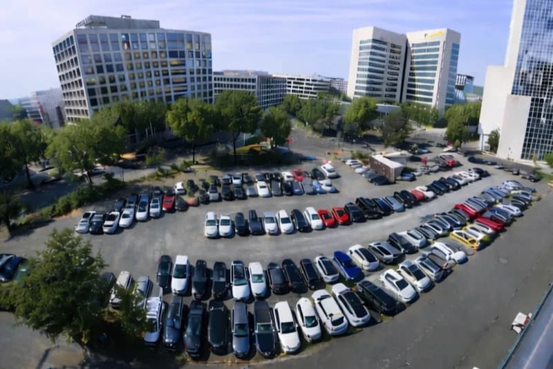 Luftaufnahme eines Parkplatzes mit in Bögen geparkten Autos, umgeben von Bürobauten und Bäumen.