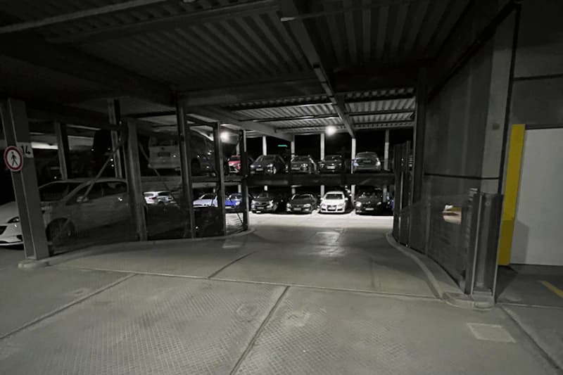 Mehrstöckiges Parkhaus mit geparkten Autos auf zwei Ebenen, beleuchteter Fahrweg und Betonboden.