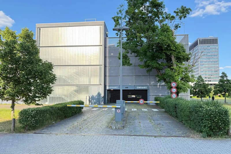 Einfahrt eines mehrstöckigen Parkhauses mit Schranke, Metallfassade, Bäumen und Verkehrsschildern.