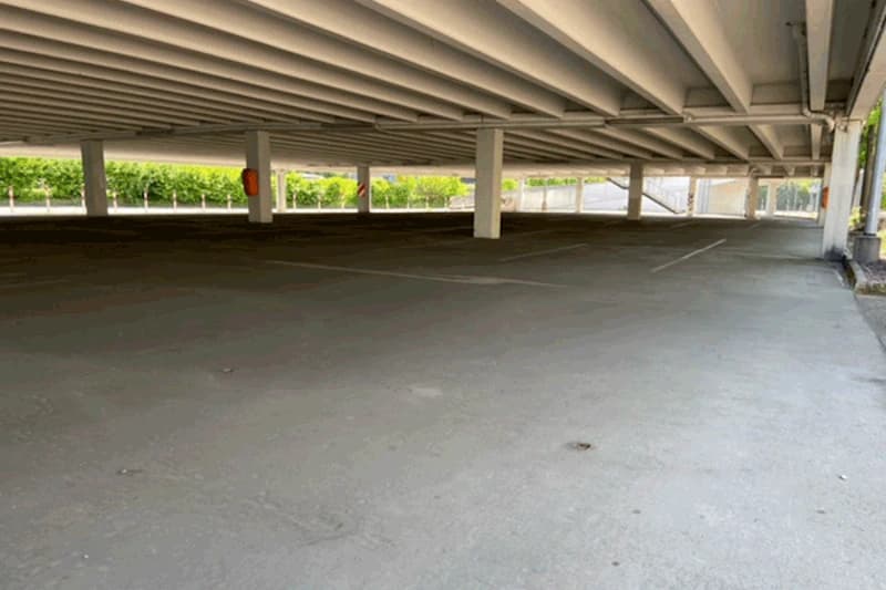 Leeres überdachtes Parkdeck mit Betonstützen, markierten Stellflächen und niedriger Betondecke, grüne Hecke im Hintergrund.
