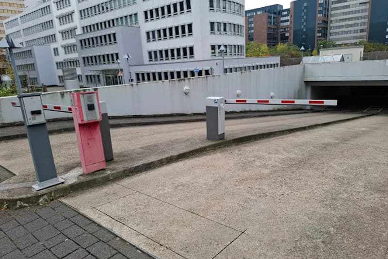 Einfahrt zur Tiefgarage mit zwei Schranken und Zahlstellen vor einer Abfahrt, im Hintergrund mehrstöckige Bürogebäude.