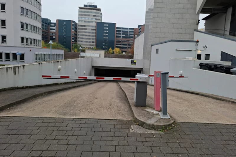Schranke über der Zufahrt zur Tiefgarage mit Ticketautomat, schräge Rampe und Bürogebäude im Hintergrund.