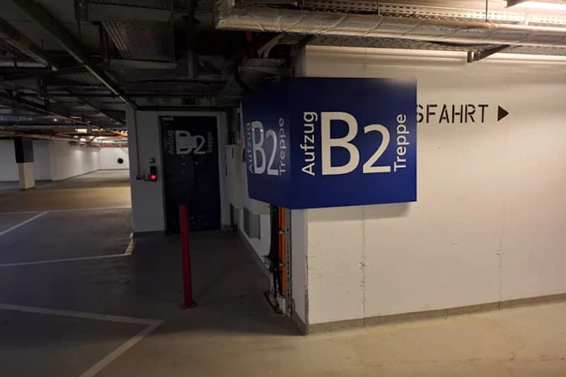 Blauer Würfel mit großer Aufschrift B2 Aufzug Treppe an einer Säule in einer Tiefgarage, weiße Wand mit Ausfahrtpfeil.