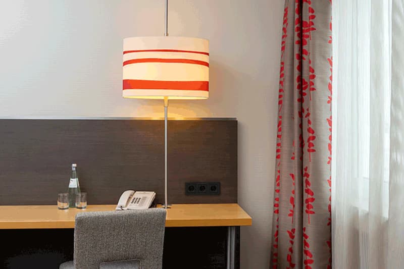 Schreibtisch mit stehender Lampe, Telefon, Wasserflasche und zwei Gläsern neben grauem Stuhl und gemustertem Vorhang.