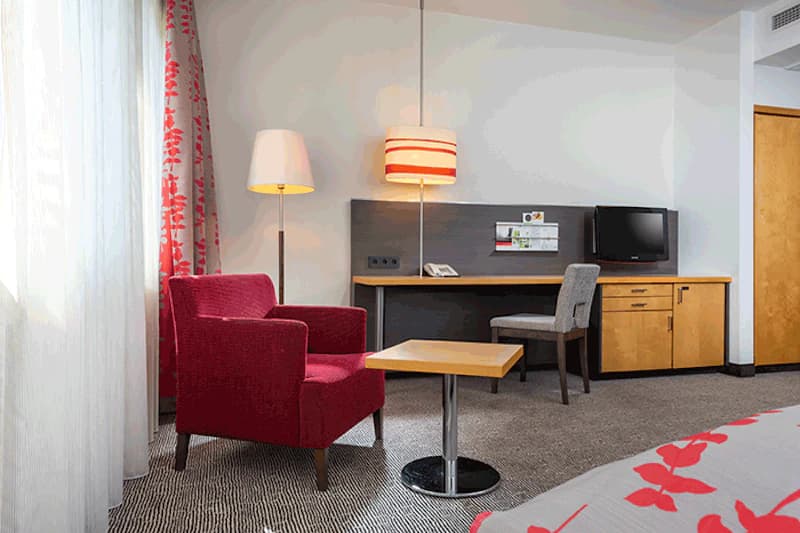 Roter Sessel neben Steh- und Hängelampe sowie kleinem Tisch vor Schreibtisch mit Stuhl, Fernseher und Schrank im Hotelzimmer.