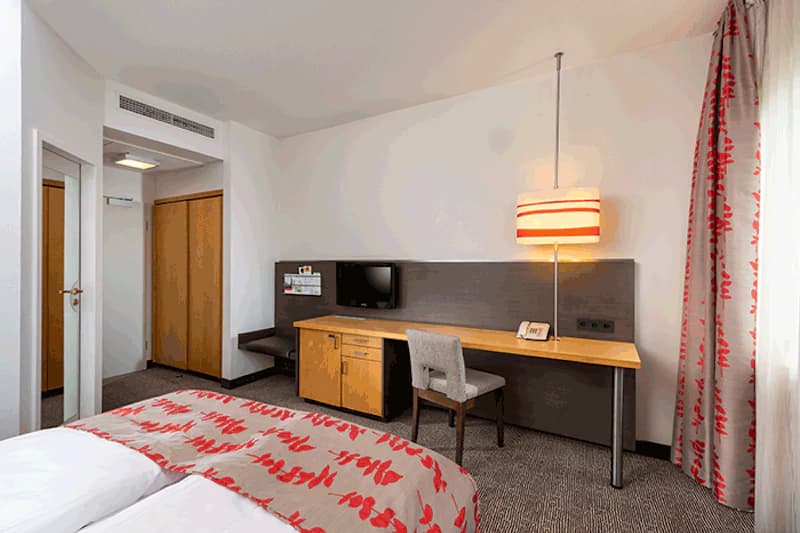 Doppelzimmer mit Doppelbett, Schreibtisch, Stuhl, Wandfernseher, Stehlampe und Kleiderschrank.