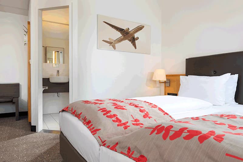 Hotelzimmer mit Doppelbett, roter Blattmusterdecke, Kopfteil, Wandlampe und offenem Bad mit Waschbecken.