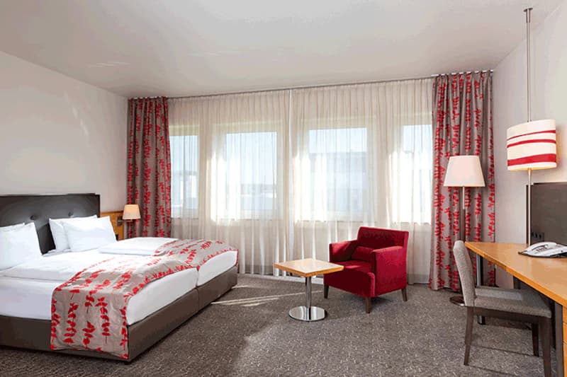 Helles Hotelzimmer mit zwei Betten, roten Vorhängen und Sessel, Schreibtisch und großen Fenstern.