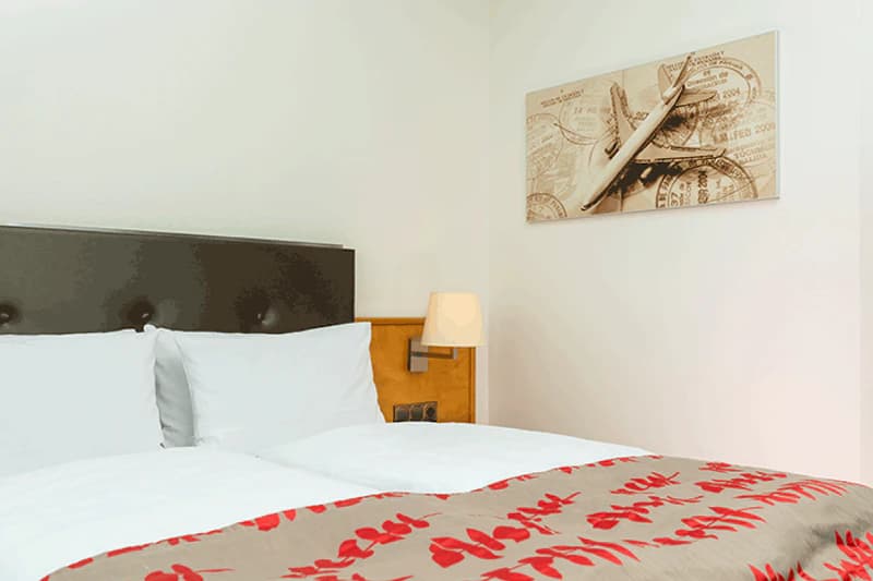 Doppelbett mit weißer Bettwäsche, dunklem Kopfteil, Nachttischlampe und Wandbild mit Flugzeug.
