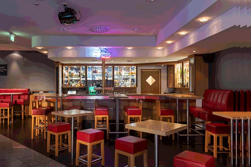 Hotelbar mit Holztheke, beleuchteten Spirituosenschränken, roten Hockern und Tischen.