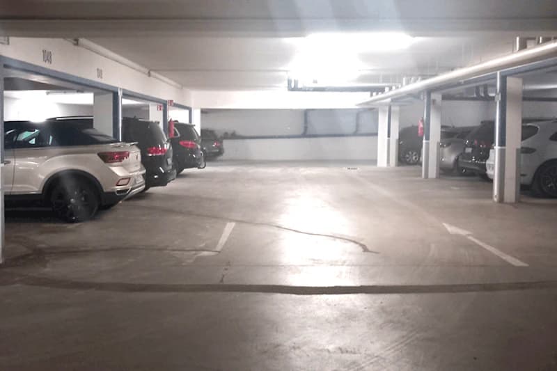 Parkdeck einer Tiefgarage mit mehreren geparkten Autos, Stützpfeilern und Deckenrohren.