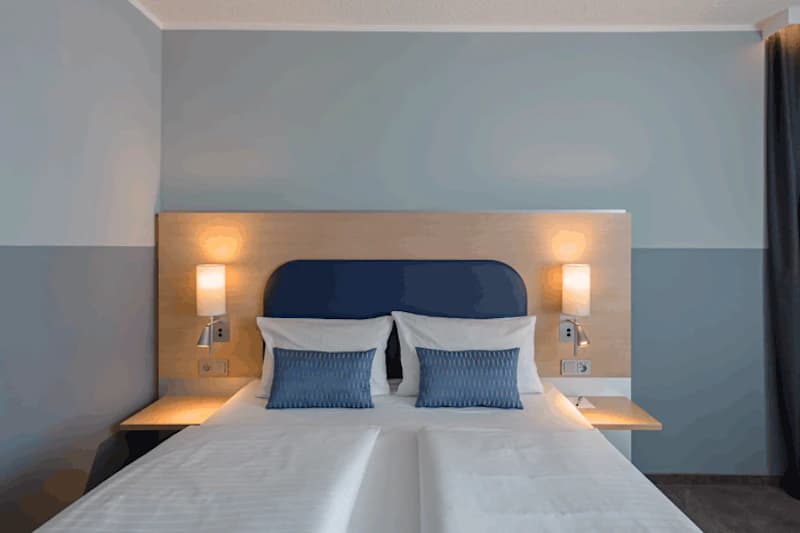 Doppelbett mit zwei Kopfkissen und blauen Zierkissen, Holzkopfteil und je eine Wandlampe auf beiden Seiten.