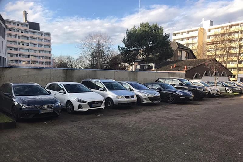 Reihe geparkter Pkw vor einer hohen Betonmauer, Wohnhäuser und Bäume im Hintergrund, bewölkter Himmel.