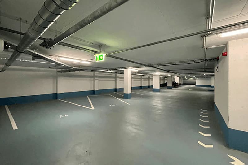 Unterirdisches Parkhaus mit leeren Stellplätzen, Stützsäulen, Lüftungsrohren und grüner Notausgangsbeleuchtung.