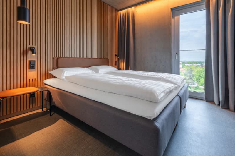 Doppelbett mit weißen Bettdecken vor Holzlattenwand, Hänge- und Leselampen, Vorhänge und Fenster mit Außenblick.