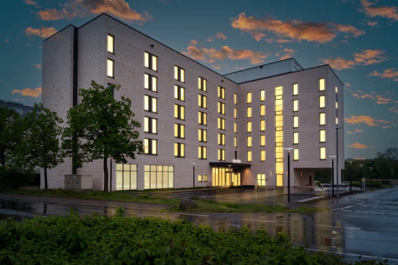 Mehrstöckiges Hotelgebäude mit leuchtenden Fensterfronten, Eingang mit Vordach und nassem Vorplatz bei Dämmerung.