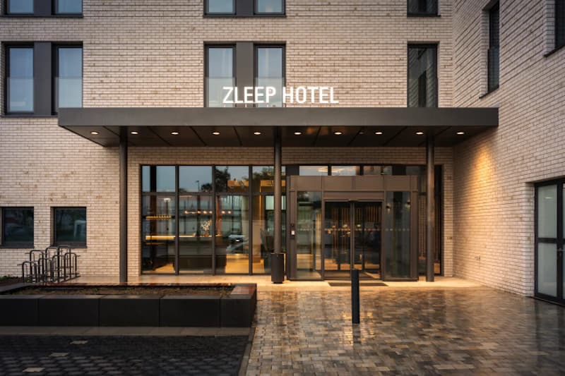 Eingangsbereich des Zleep Hotel mit Glas-Eingangstüren, Vordach und heller Backsteinfassade bei Abendlicht.