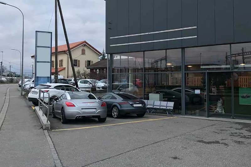 Parkplätze vor Glasfassade mit mehreren geparkten Autos, Gehweg und leerem Werbepylon bei bedecktem Himmel.