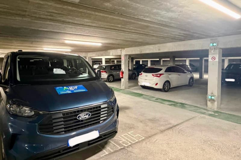 Überdachte Parkhalle mit mehreren geparkten Autos, vorn ein blauer Ford mit Agir-Aufkleber, Betonstützen und Deckenbeleuchtung.