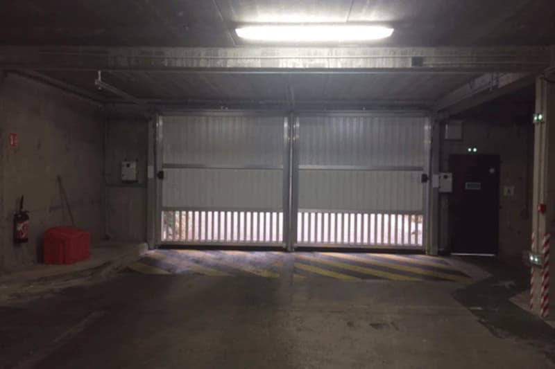 Teilweise geschlossene Metalltore einer Tiefgarage mit Lichtspalt unten, Betonwände, gelb-schwarze Bodenmarkierung und Feuerlöscher links.