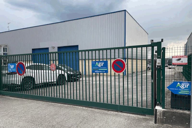 Grünes Metall-Schiebetor mit Parkverbotsschildern und Agir-Schild, dahinter weißes Auto und Halle mit blauen Toren.