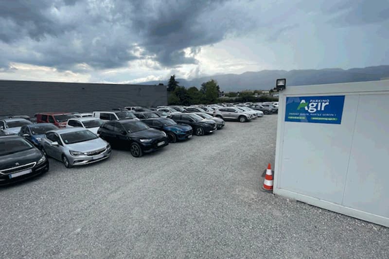 Grosser Kiesparkplatz mit Reihen geparkter Autos und weissem Container mit Parking Agir Schild unter bewölktem Himmel.