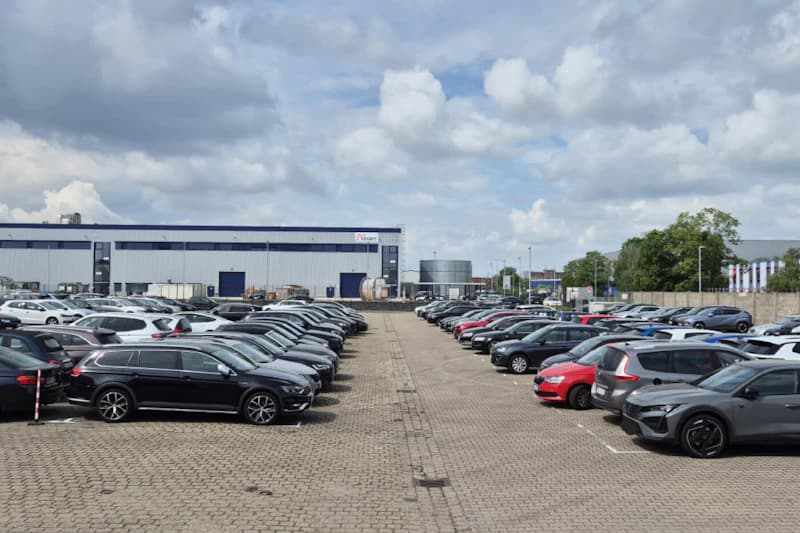 Großer Außenparkplatz mit Reihen geparkter Autos vor einem Industriegebäude und bewölktem Himmel.