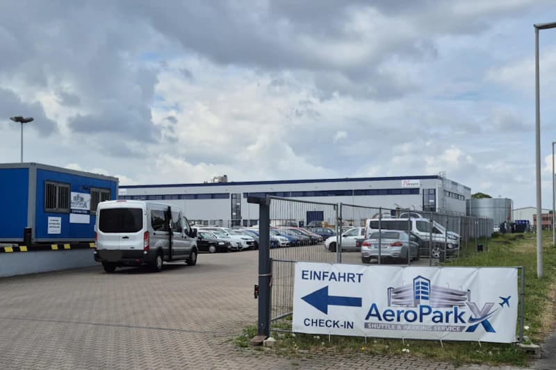 Eingezäunter Parkplatz mit parkenden Autos und Vans, Einfahrt-Banner von AeroParkX und Industriegebäude im Hintergrund.