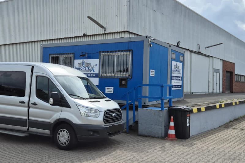 Silberner Shuttlebus neben blauem Bürocontainer mit AeroParkX Schild vor einer Lagerhalle.