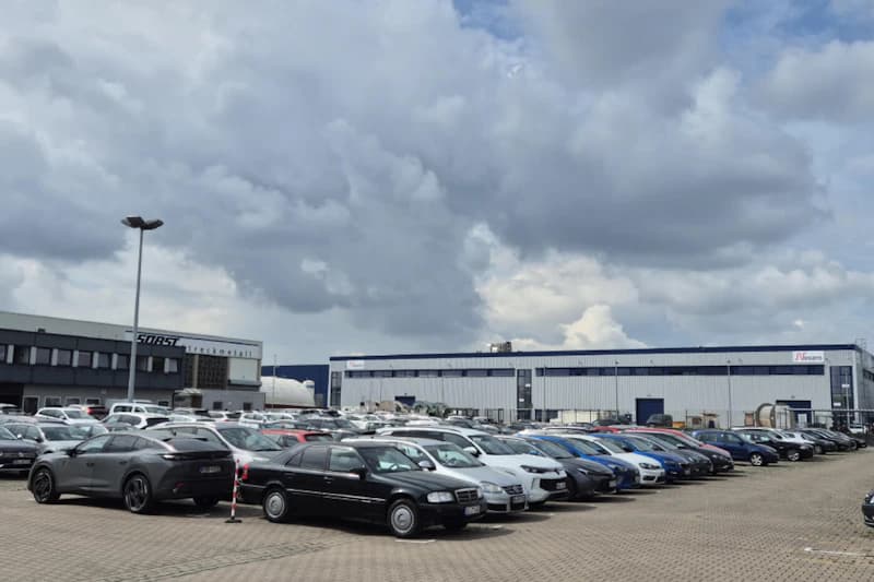 Großer Parkplatz mit vielen geparkten Autos auf gepflasterter Fläche vor Industriehallen und bewölktem Himmel.