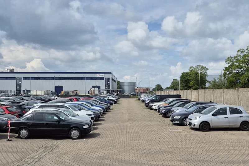 Außenparkplatz mit zwei Reihen geparkter Autos, Fahrweg in der Mitte, Industriegebäude und Betonmauer im Hintergrund.