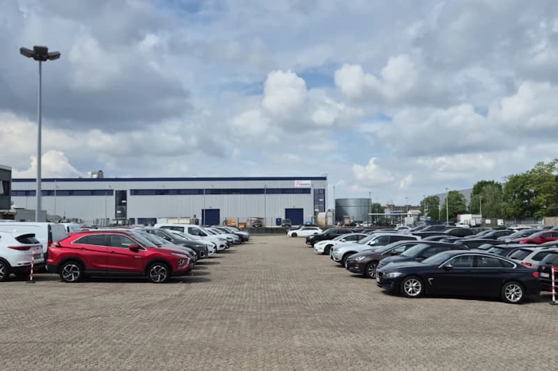 Großer Außenparkplatz mit zahlreichen in Reihen geparkten Autos und einer Lagerhalle im Hintergrund.