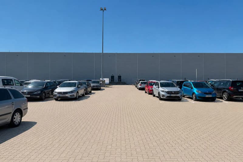 Gepflasterter Parkplatz mit geparkten Autos an beiden Seiten, mittigem Lichtmast und großer Hallenfassade im Hintergrund.