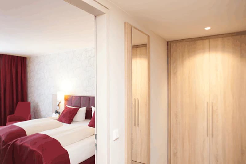Hotelzimmer mit zwei Betten, roten Decken und Kissen, Nachttischlampe, roten Vorhängen und hellem Holzkleiderschrank mit Spiegel.