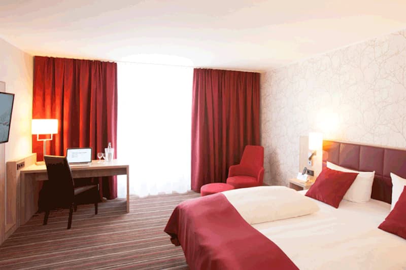 Hotelzimmer mit Doppelbett und roter Bettdecke, Schreibtisch mit Laptop, rotem Sessel und bodentiefen Vorhängen.