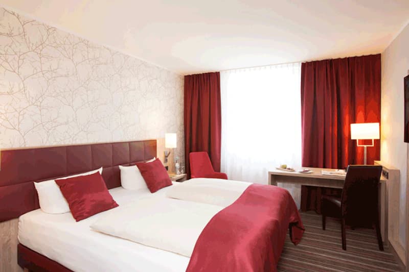 Hotelzimmer mit zwei Einzelbetten, roter Bettwäsche und Kissen, Schreibtisch, Stuhl und Vorhang.
