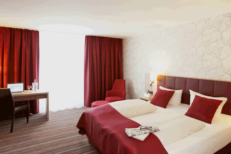 Hotelzimmer mit Doppelbett, roten Vorhängen, Sessel, Schreibtisch und Zeitung auf dem Bett.