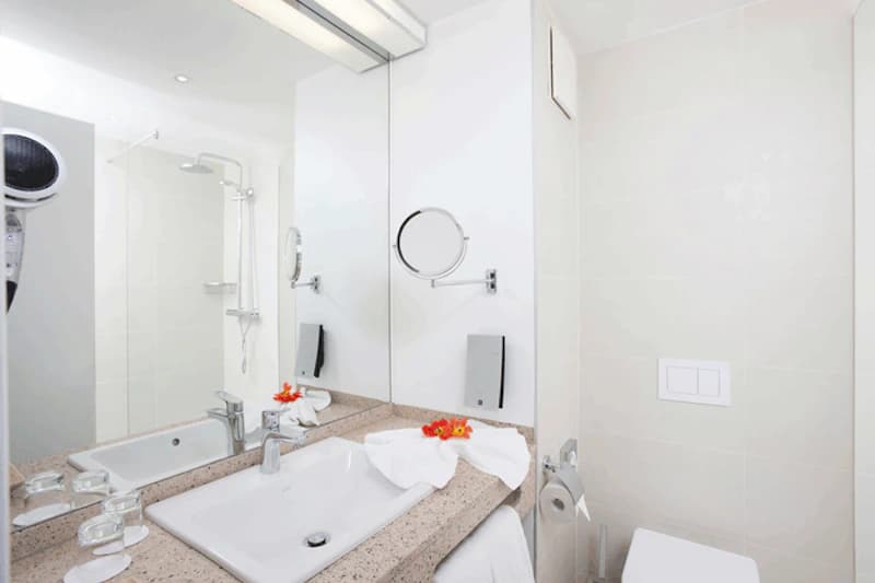 Helles Hotelbadezimmer mit Waschbecken, großem Spiegel, Dusche, Toilette und Handtuch mit Blumen.