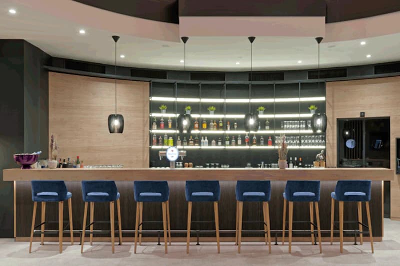 Hotelbar mit langer Holztheke, sieben blauen Barhockern und beleuchteten Regalen mit Flaschen.