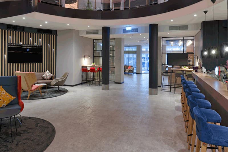 Offene Hotellobby mit Bar und blauen Hockern, Sitzgruppen, zwei schwarzen Säulen und Glas-Eingangstüren.