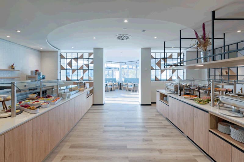 Heller Frühstücksraum mit zwei langen Buffetzeilen, Servierflächen und Sitzbereich im Hintergrund.