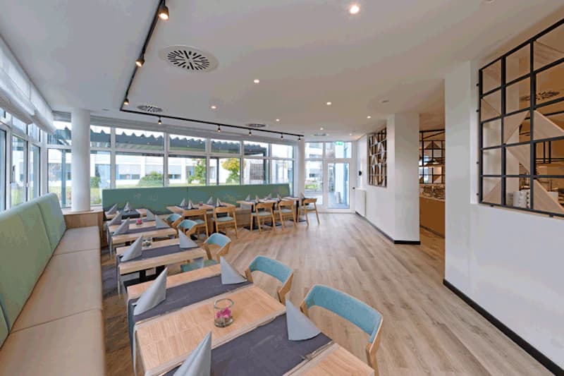Heller Hotel-Speiseraum mit Holzboden, eingedeckten Tischen, grünen Polsterbänken und großen Fenstern.