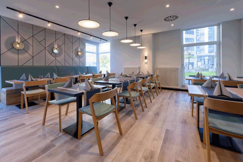 Heller Hotelrestaurant mit Tischen, Holzstühlen, gefalteten Servietten und Hängeleuchten.