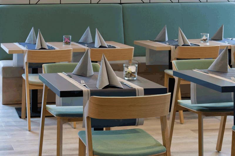 Tischreihe in Hotelrestaurant mit gefalteten Servietten, Holzstühlen und grüner Polsterbank.