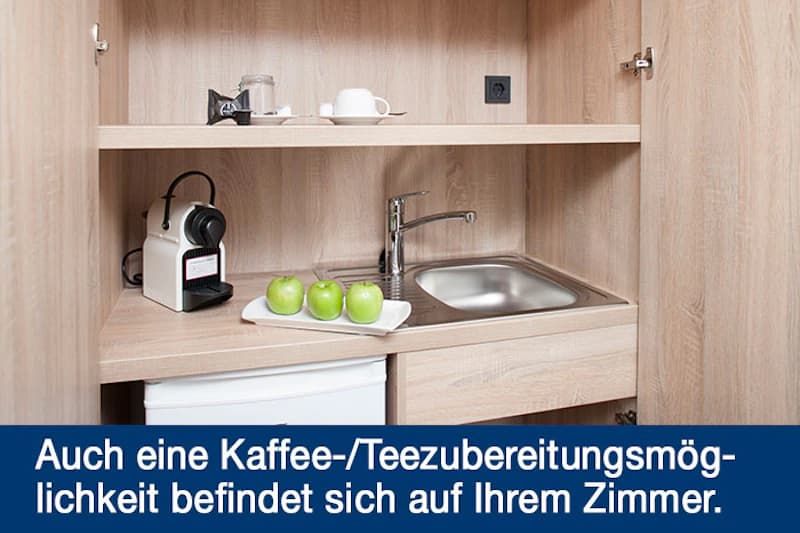 Küchenzeile im Hotelzimmer mit Kaffeemaschine, drei grünen Äpfeln auf Teller, Spüle, Tassen und kleinem Kühlschrank.