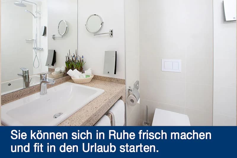 Hotelbadezimmer mit Waschbecken, Spiegel, Duschbereich, Toilette und Ablage mit Pflegeartikeln.