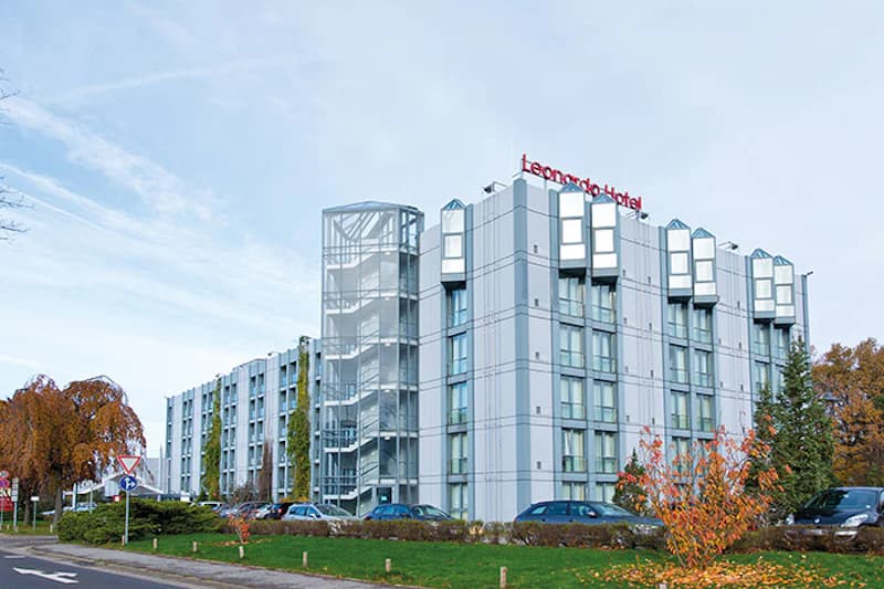 Mehrstöckiges Hotelgebäude mit außenliegender Treppe, geparkten Autos und herbstlichen Bäumen, Schriftzug Leonardo Hotel auf dem Dach.