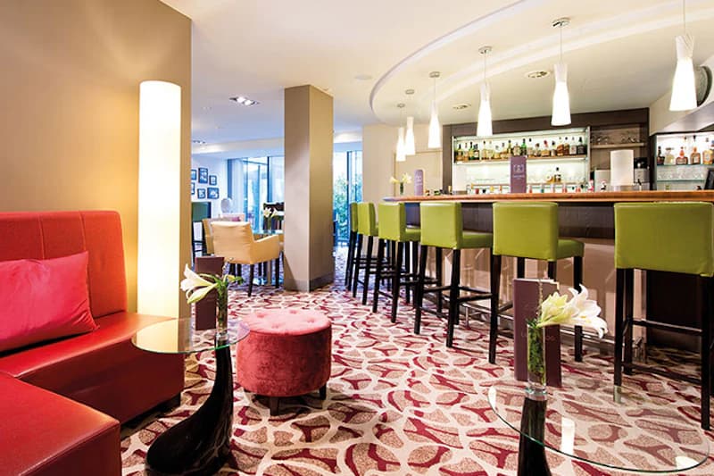 Hotelbar mit Tresen und grünen Barhockern, roter Sitzecke, gemustertem Teppich und Tischvasen mit Blumen.