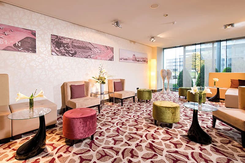 Lounge mit Sitzgruppen, bunten Samthockern, Glastischen, gemustertem Teppich und bodentiefem Fenster.